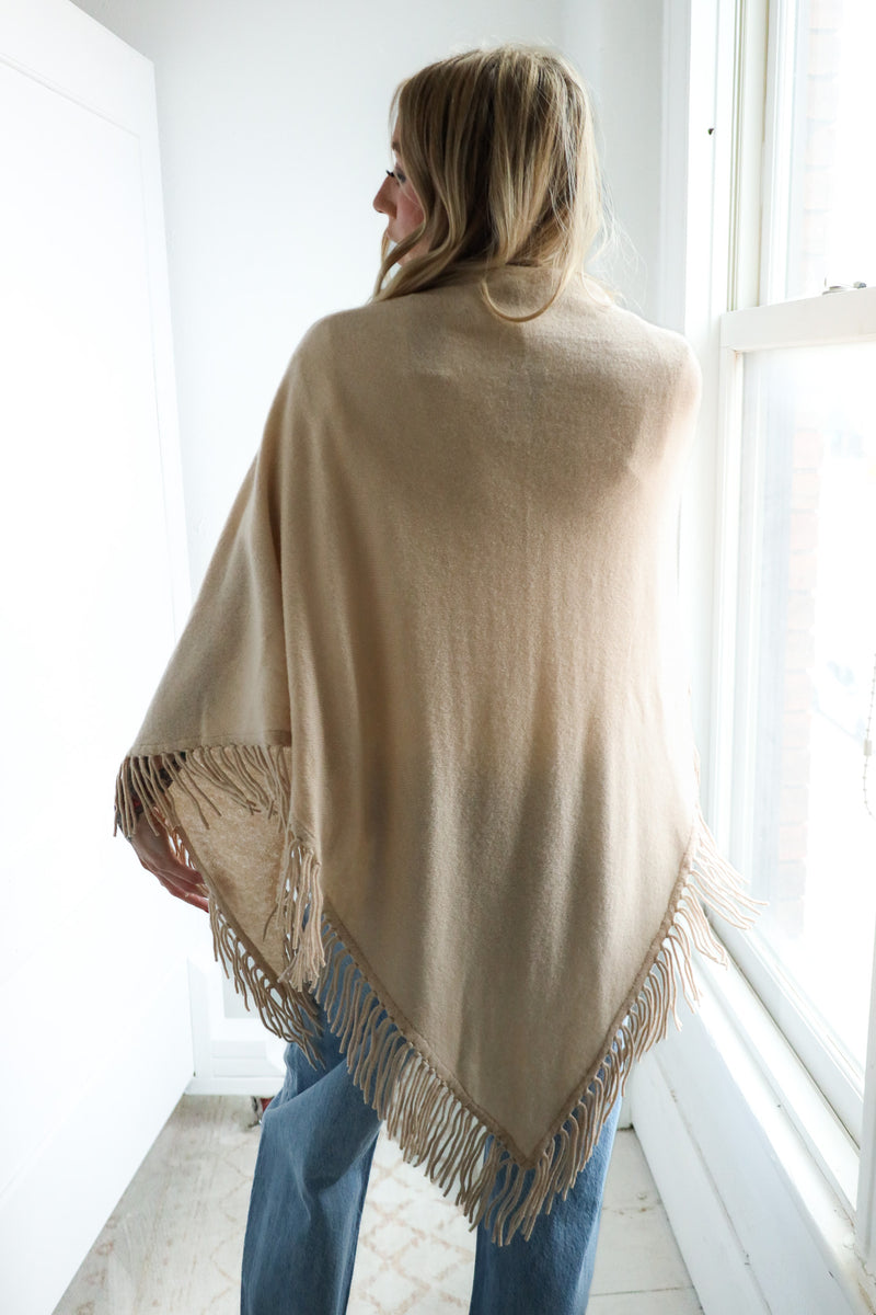 Woman wearing a beige poncho with fringe details in a bright room.