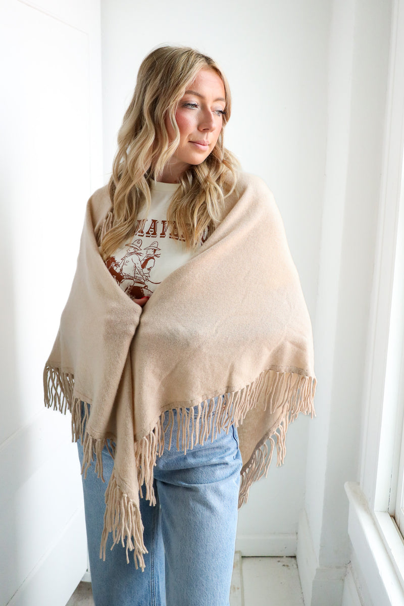 Woman wearing a beige shawl over a graphic t-shirt and jeans indoors.
