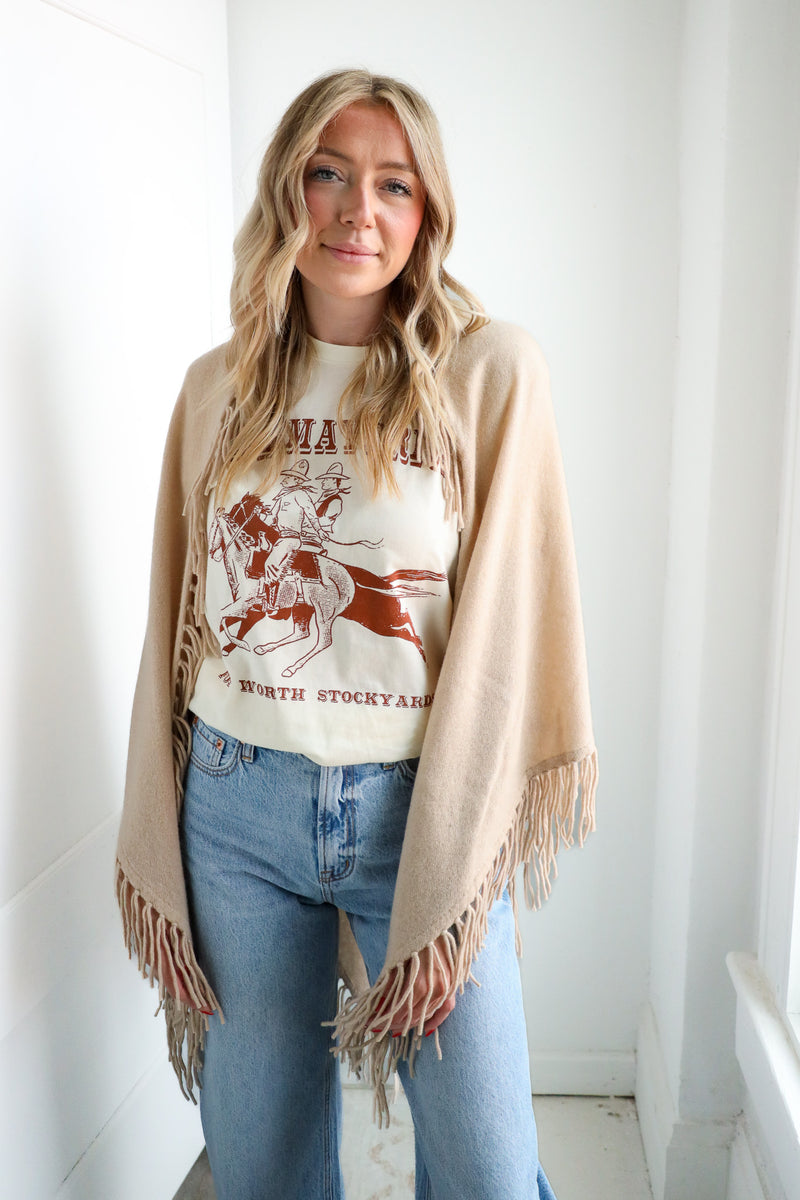 Woman wearing a graphic t-shirt, blue jeans, and a beige cardigan with fringe.