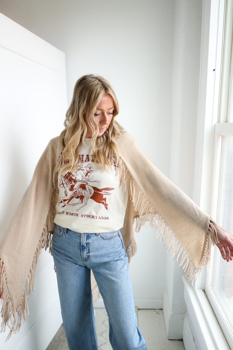 Woman wearing a white graphic t-shirt, beige cardigan, and blue jeans indoors.
