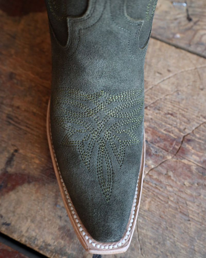 Close-up of a green suede boot with intricate stitching on a wooden floor.