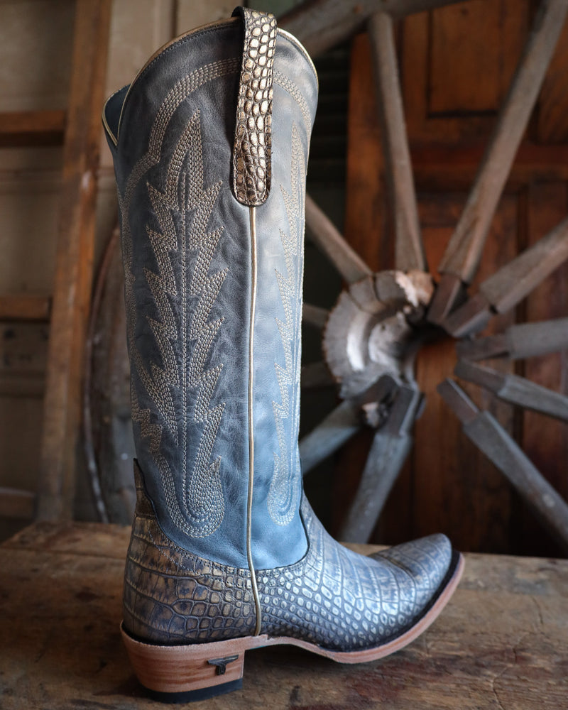 Blue cowboy boot with intricate design and studded detail in front of a wooden wagon wheel.