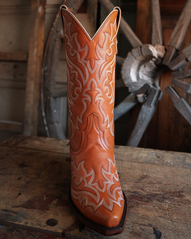 Brown cowboy boot with intricate white embroidery on a wooden surface with a rustic background.