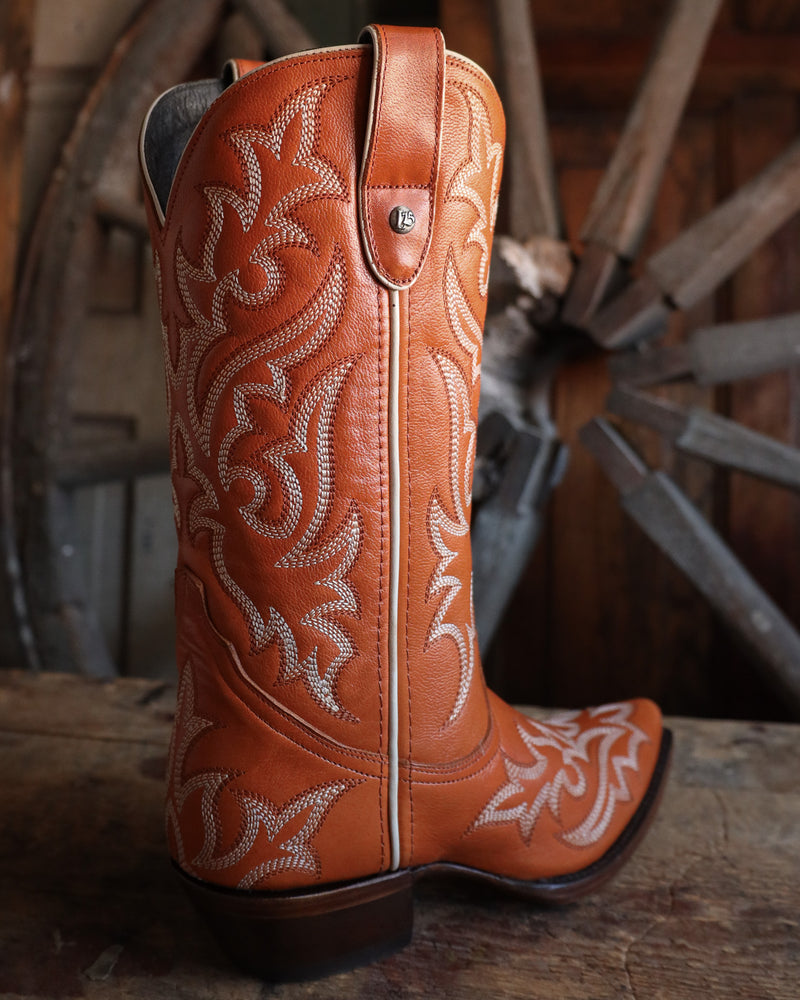 Brown cowboy boot with intricate white embroidery on a wooden surface with a wheel in the background