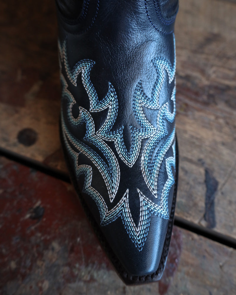 Navy blue leather boot with intricate white embroidery on a wooden floor.