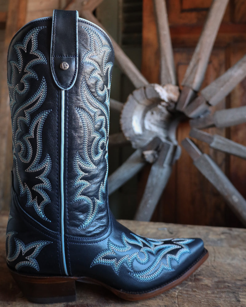 Navy blue cowboy boot with blue embroidery in front of a wooden wagon wheel.