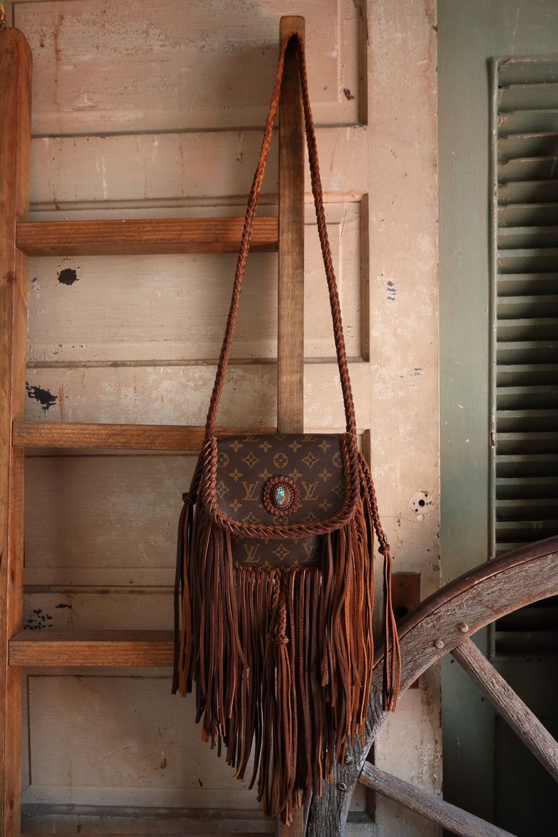 Brown leather handbag with fringes hanging on a wooden ladder against a rustic wall.