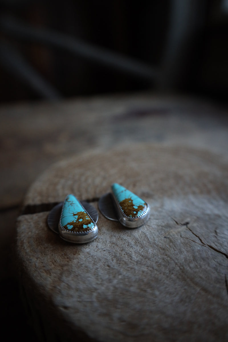 Teardrop-shaped earrings with turquoise tops and gold accents on a wooden surface.