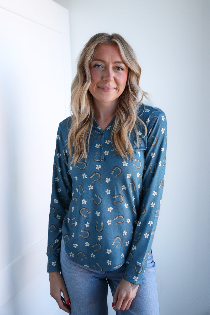Woman wearing a blue long-sleeve shirt with floral and horseshoe pattern against a white wall.