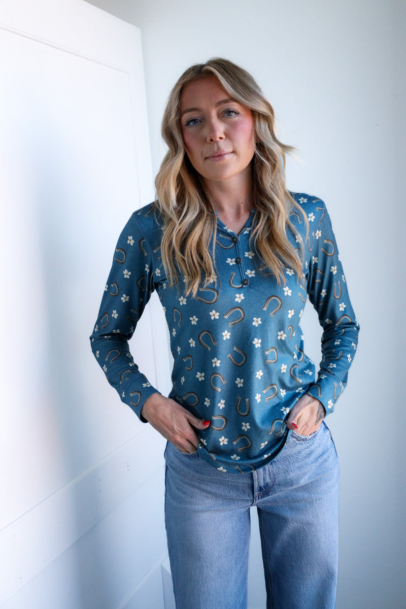 Woman wearing a blue patterned long-sleeve shirt and jeans against a white wall.