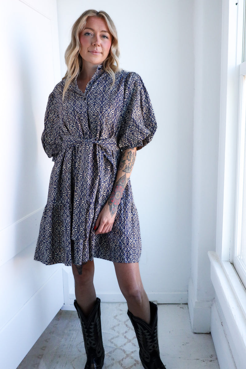Woman wearing a patterned dress standing in a bright room with white walls and a window.