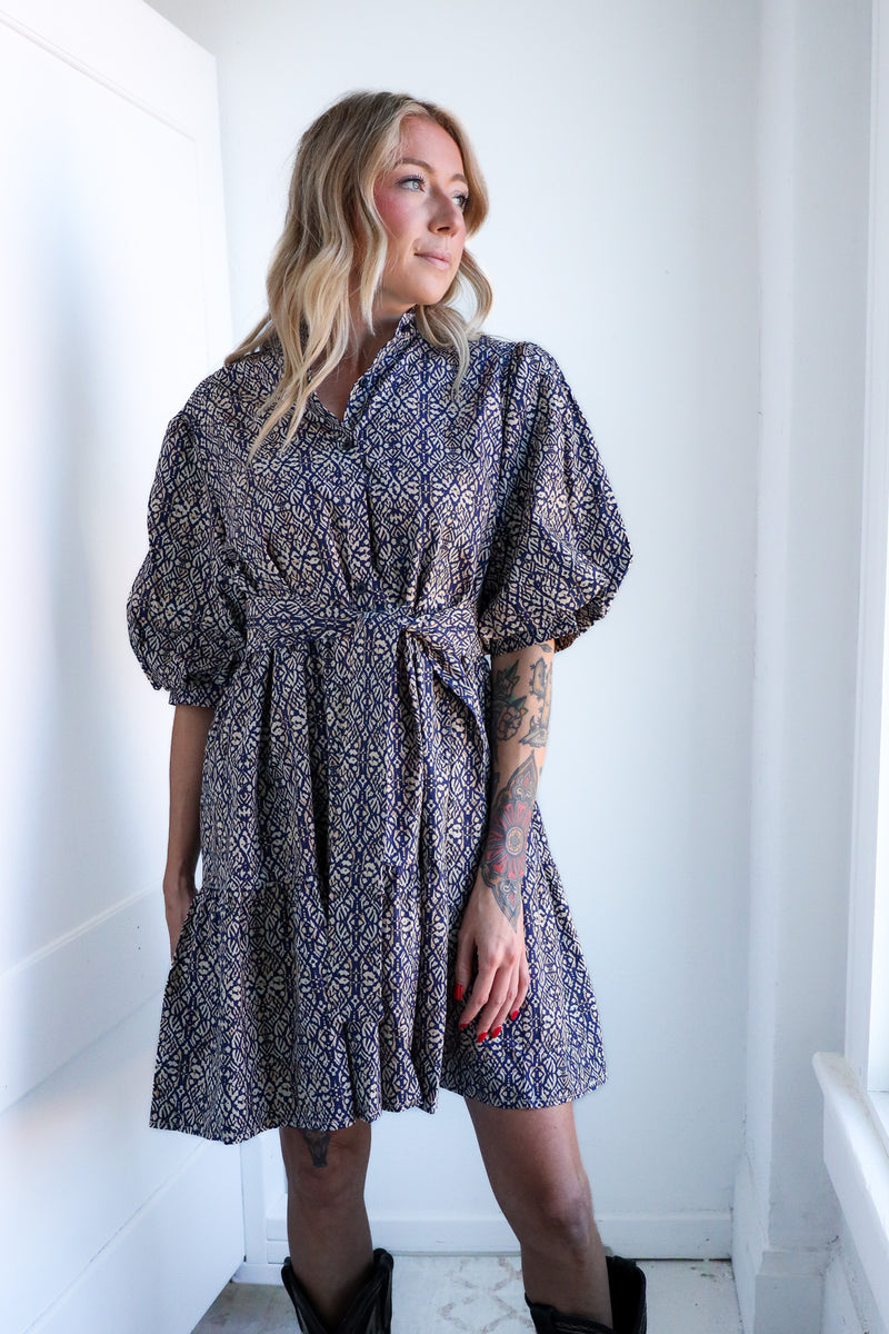 Woman wearing a patterned dress standing in a bright room with white walls.