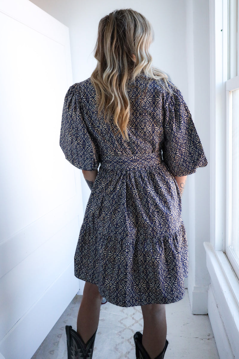 Person wearing a patterned dress standing in a bright room with white walls and a window.