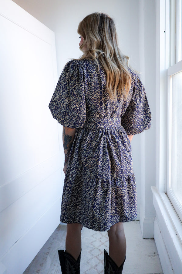 Woman wearing a patterned dress standing in a bright room with white walls and a window.