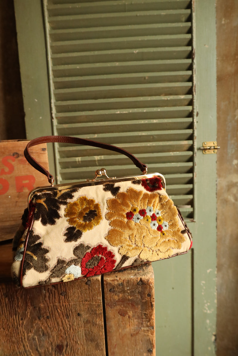 Vintage inspired style purse. Plush multicolor floral design on cream back ground. Gold kisslock hardware. Brown leather handle and base with foot studs. Fuchsia color satin lining