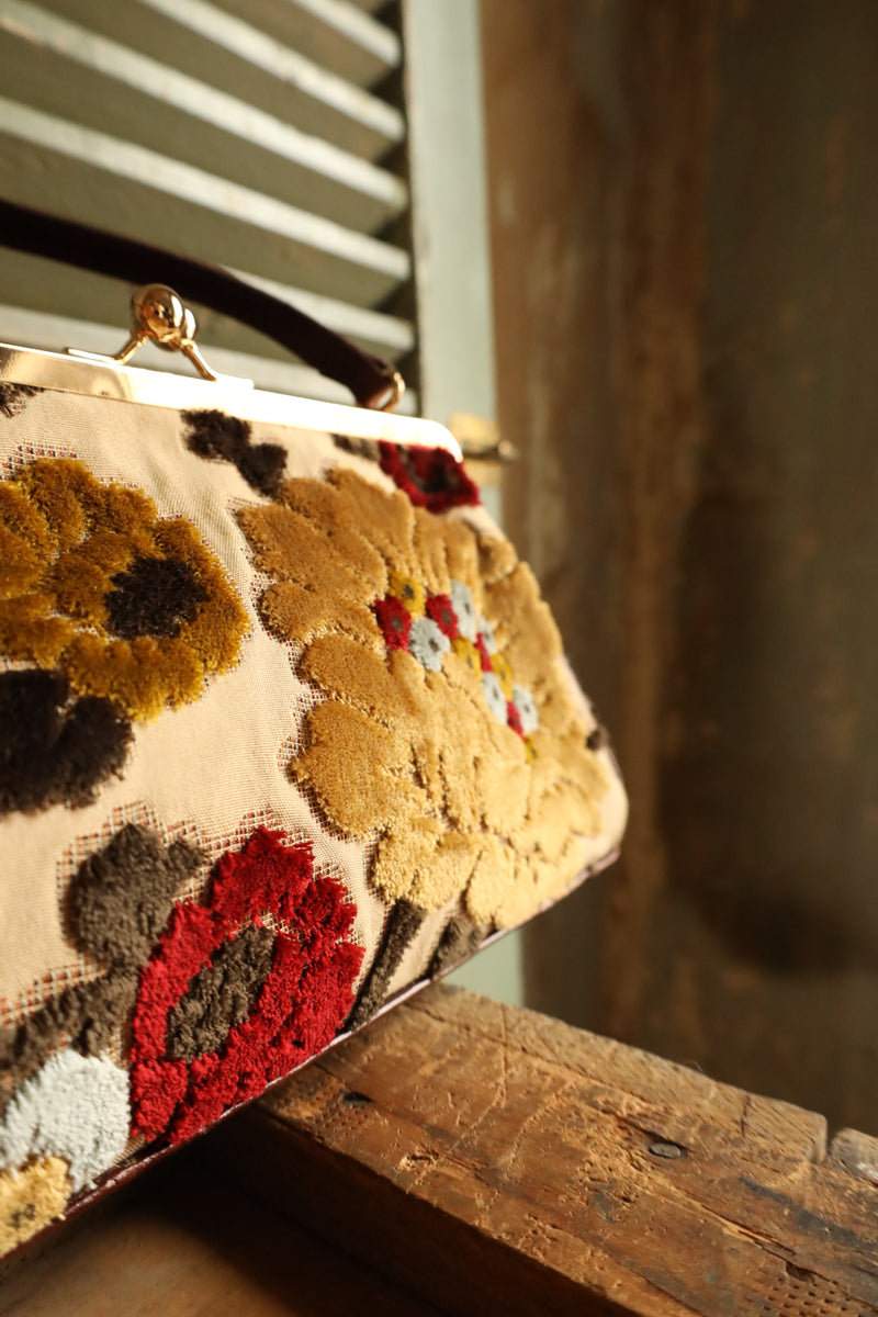 Vintage inspired style purse. Plush multicolor floral design on cream back ground. Gold kisslock hardware. Brown leather handle and base with foot studs. Fuchsia color satin lining