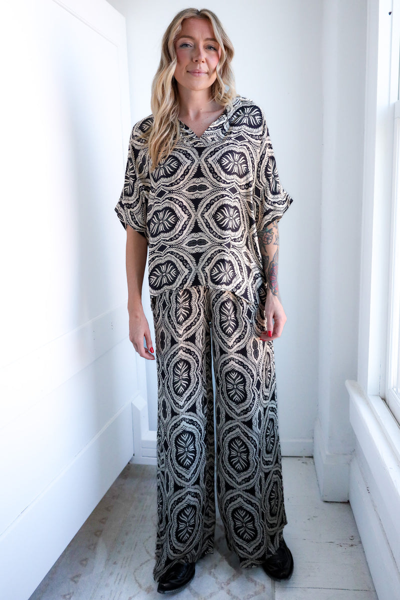 Woman wearing a patterned outfit standing in a bright room with white walls and a window.