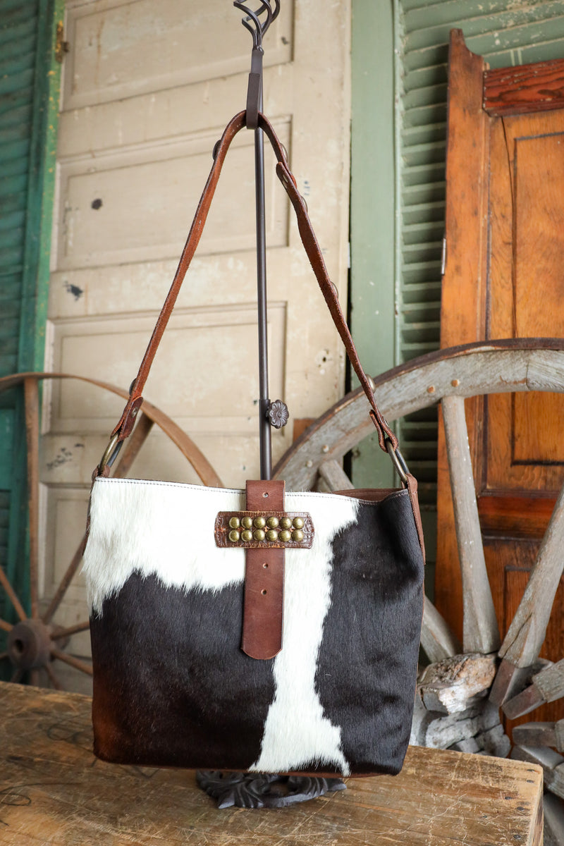Black and white cowhide handbag with brown leather strap and studs, hanging against a rustic wooden background.