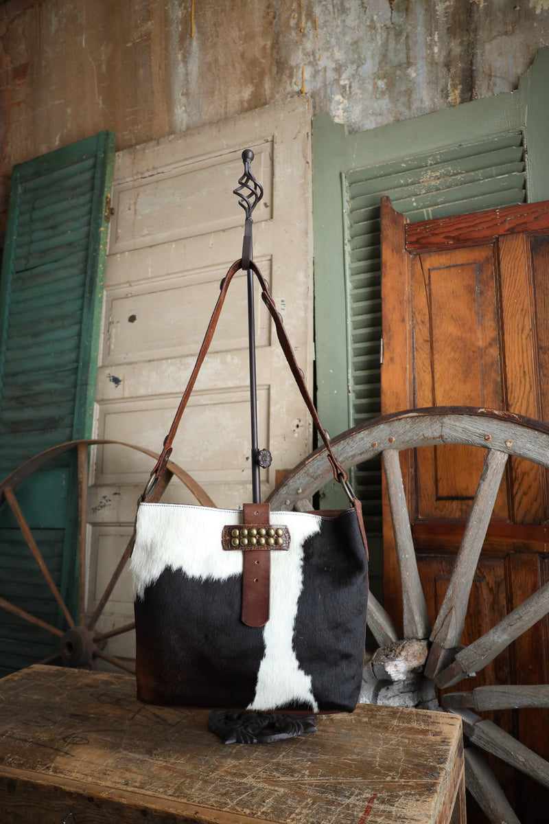 Black and white cowhide handbag with brown leather strap on a wooden surface with rustic background