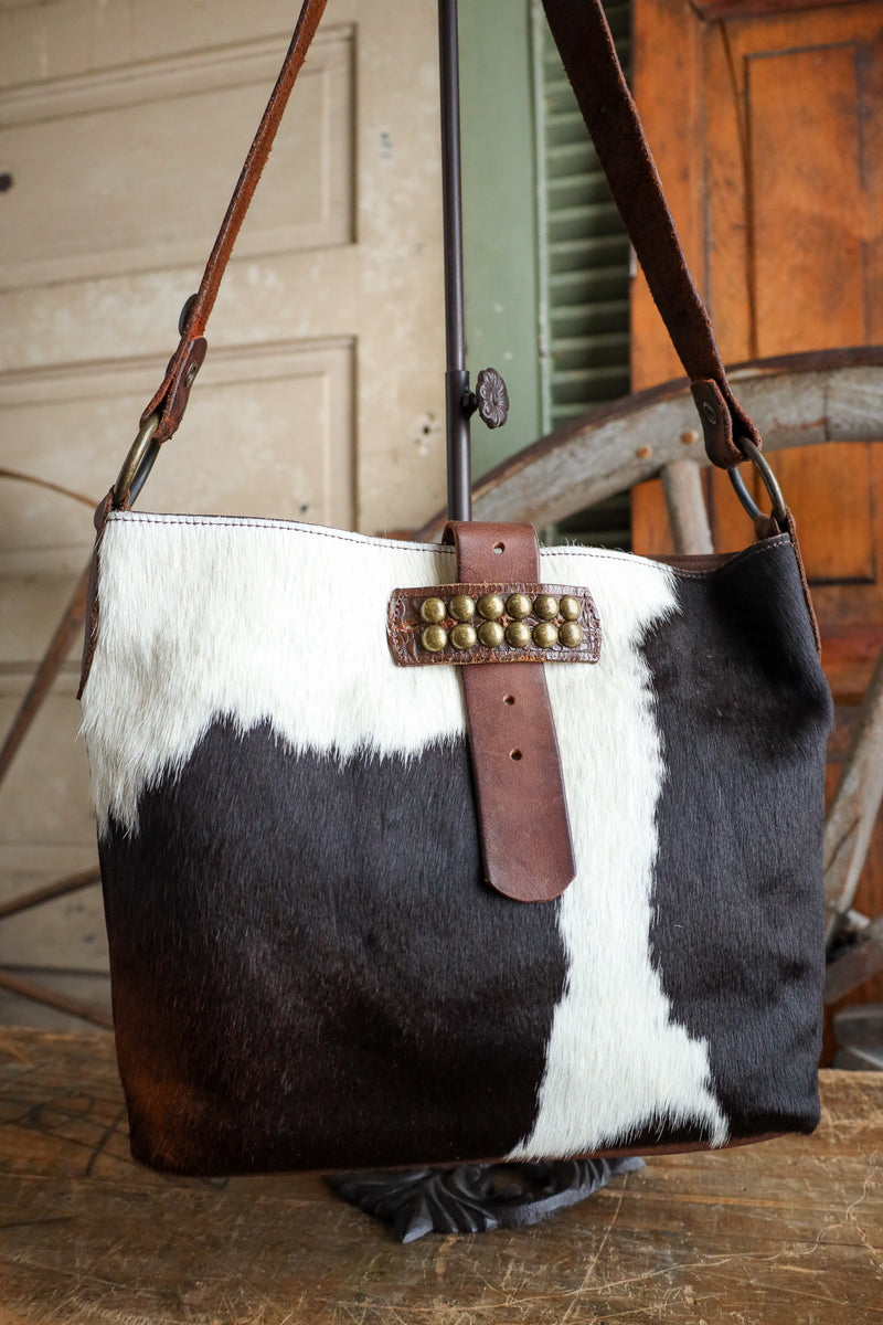 Black and white cowhide handbag with brown leather strap on a wooden surface.