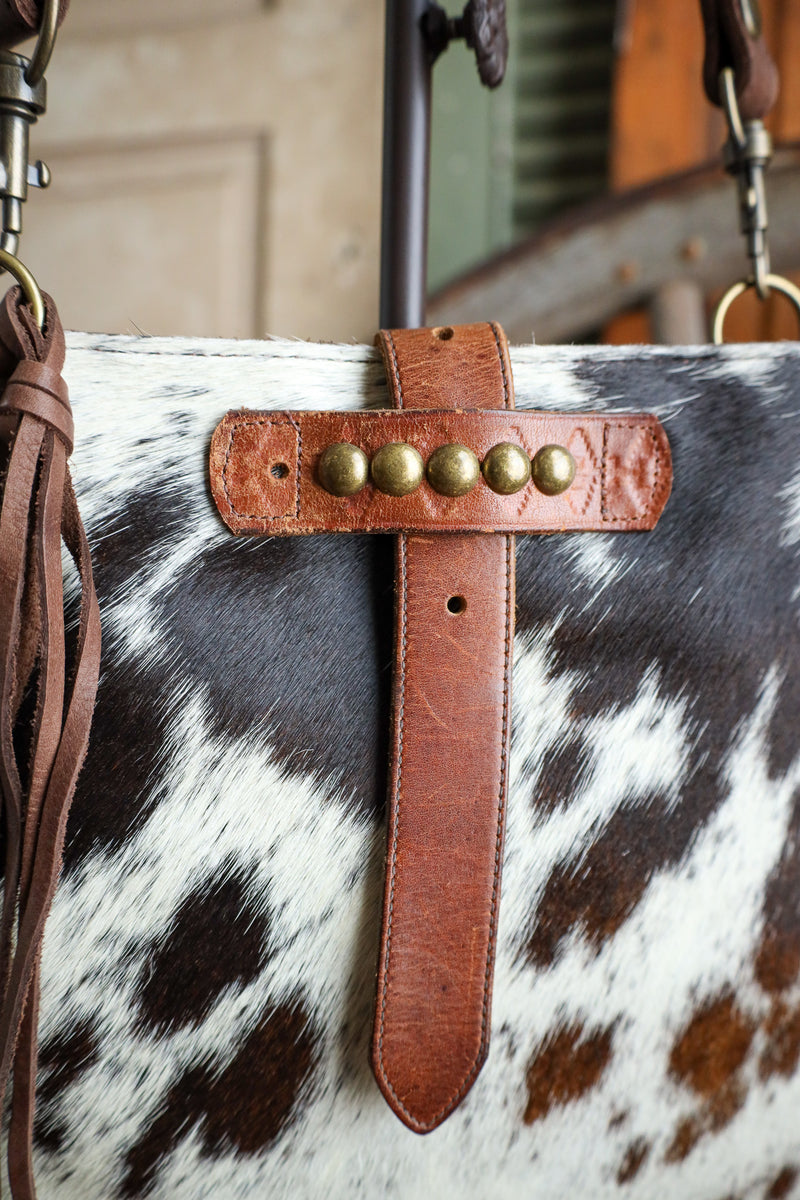 Close-up of a cowhide bag with a brown leather strap featuring brass studs.