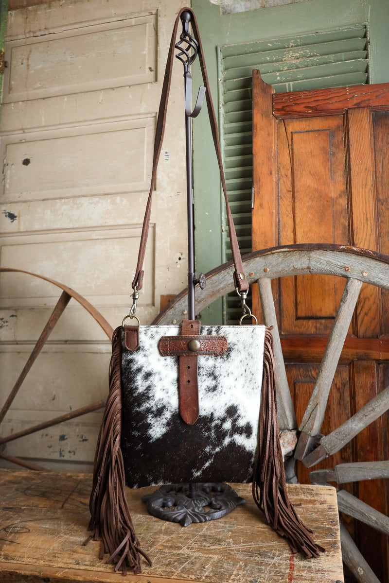 Cowhide leather handbag with fringe and cross detail on a wooden surface.