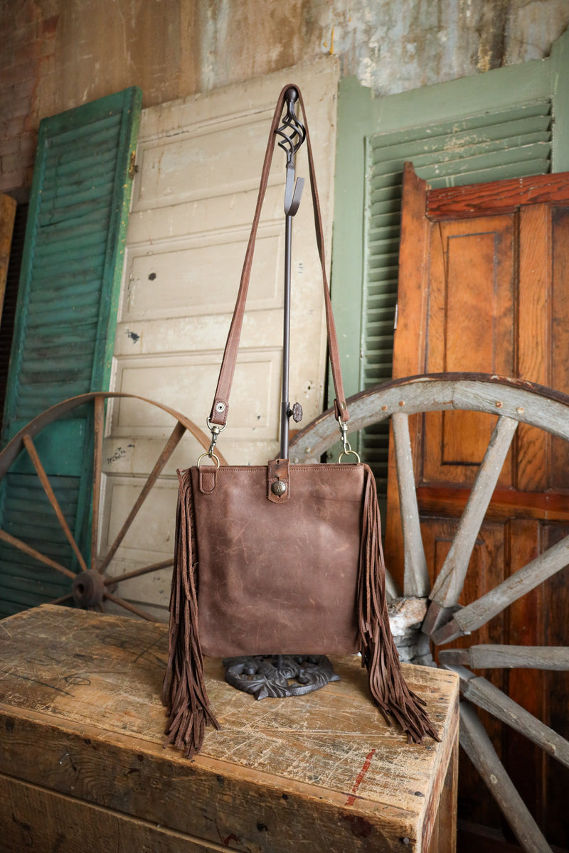 Brown leather handbag with fringe on a wooden surface in front of rustic wooden doors and a wagon wheel.