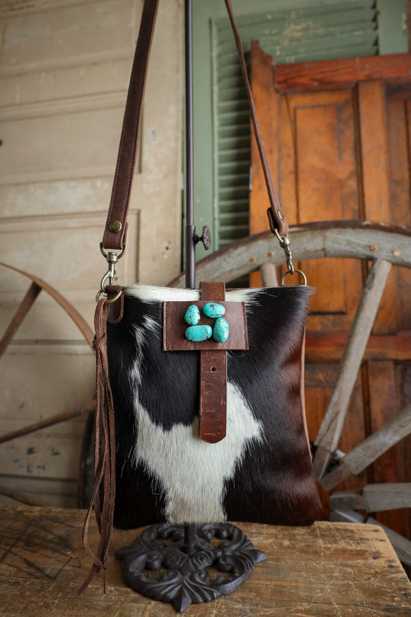 Cowhide handbag with turquoise stones and brown leather strap on a rustic wooden surface.