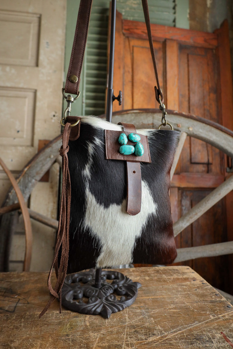 Cowhide bag with leather strap and turquoise accents on a wooden surface.