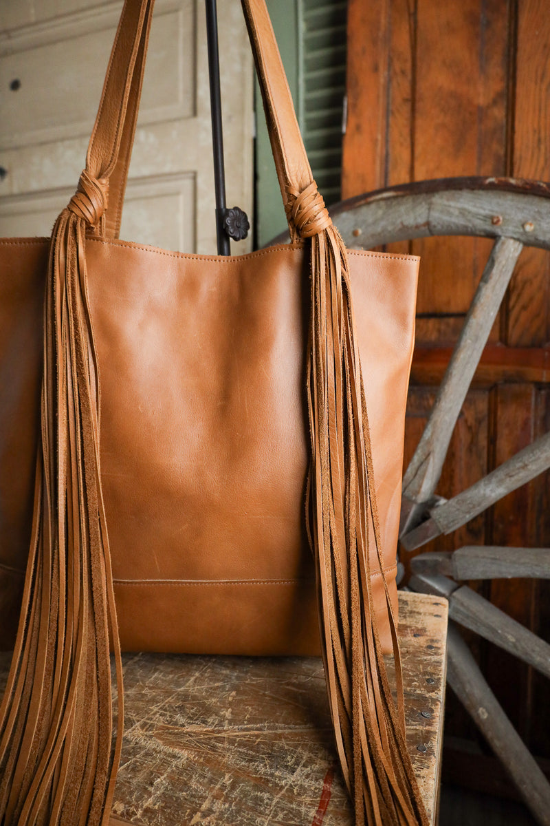 Brown leather tote bag with fringes in front of a wooden wheel