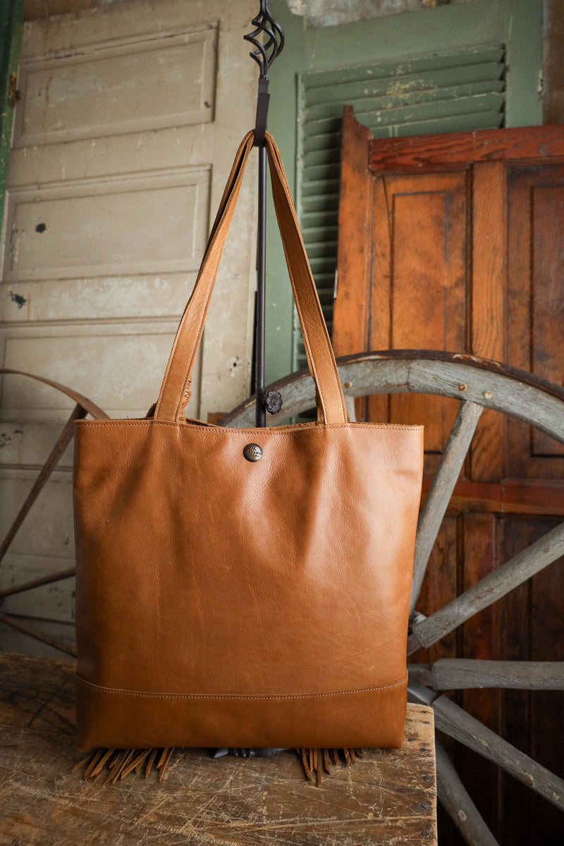 Brown leather tote bag with fringe hanging on a wooden wheel against a rustic background