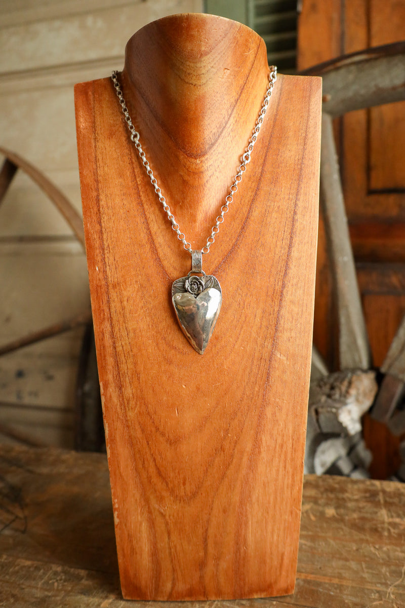 Silver heart-shaped pendant on a chain displayed on a wooden stand.
