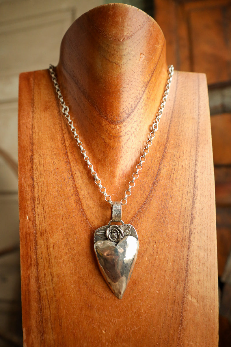 Silver heart-shaped pendant on a chain displayed on a wooden stand.