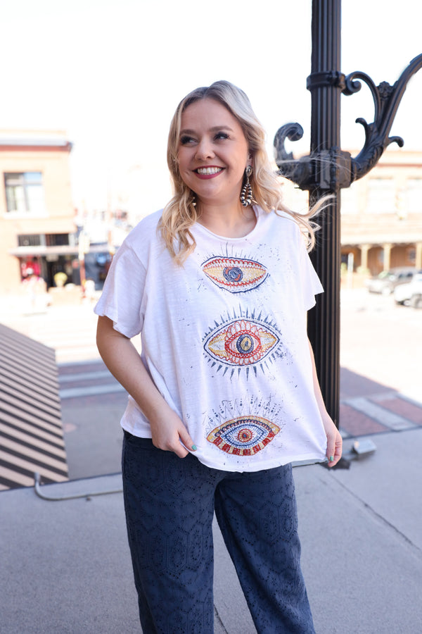 Woman wearing a white t-shirt featuring embroidered three eyes in cool and warm tones, with a playful blue splatter paint detail. Stand out from the crowd and add a touch of eccentricity to your wardrobe with this must-have tee!