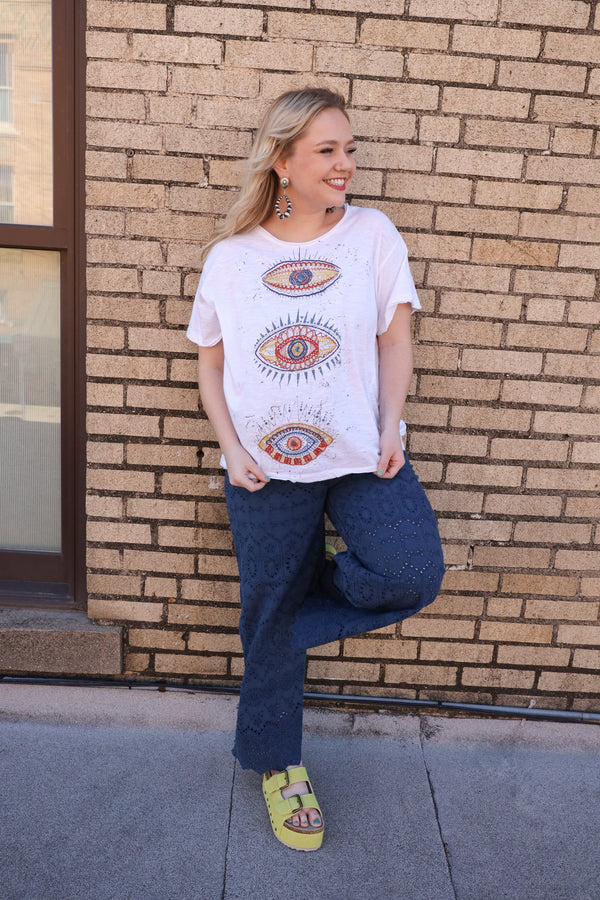 Woman wearing a white t-shirt featuring embroidered three eyes in cool and warm tones, with a playful blue splatter paint detail. Stand out from the crowd and add a touch of eccentricity to your wardrobe with this must-have tee!
