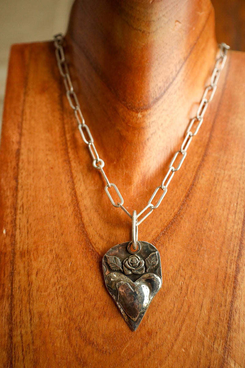 Silver chain necklace with a heart-shaped pendant featuring a rose design on a wooden mannequin.