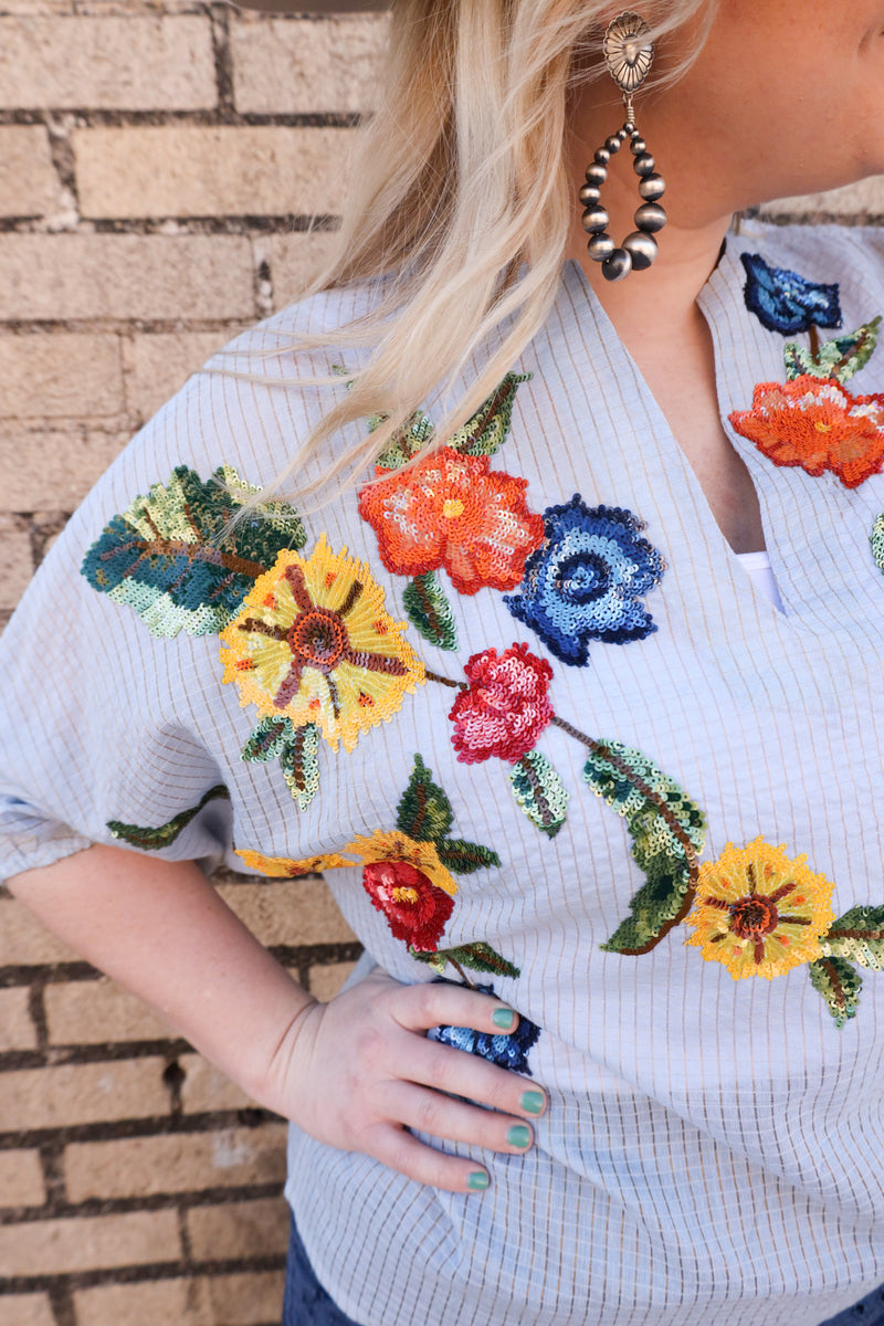 Woman wearing oversized blouse features a stunning blue checker print, a flirty v-neck, and chic quarter sleeve cinch detail. Adorned with multi-colored sequin flowers, this blouse will elevate any outfit with a touch of whimsy.