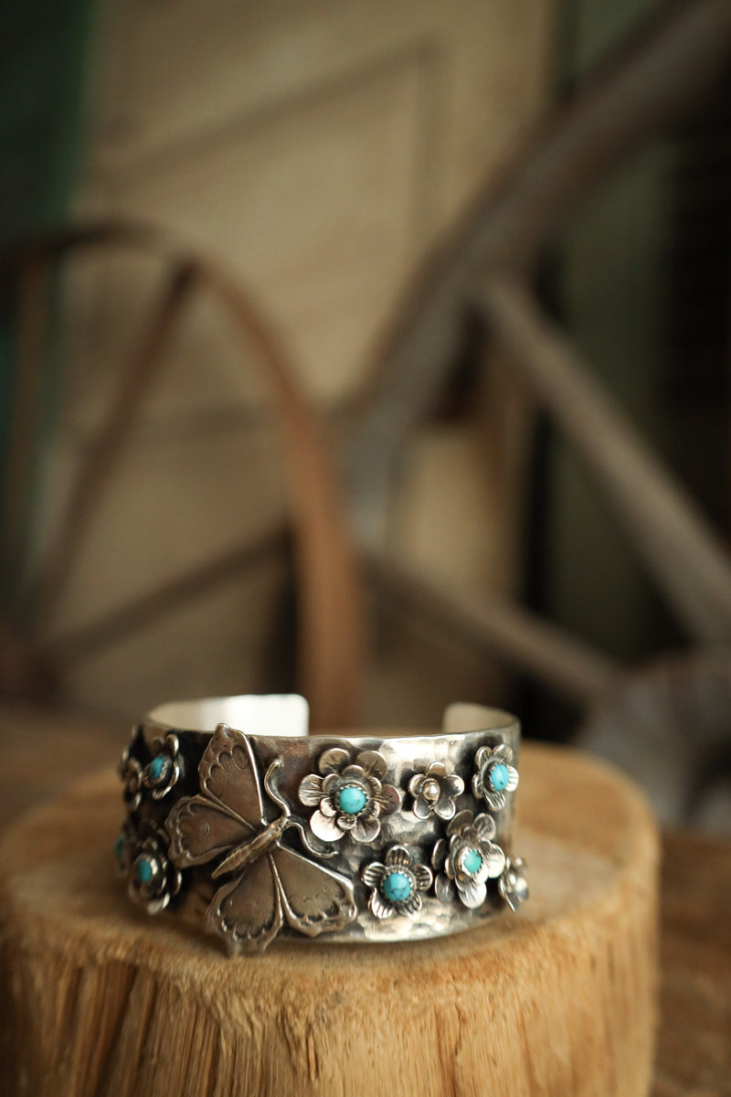 Silver cuff bracelet with floral designs and turquoise stones on a wooden block.