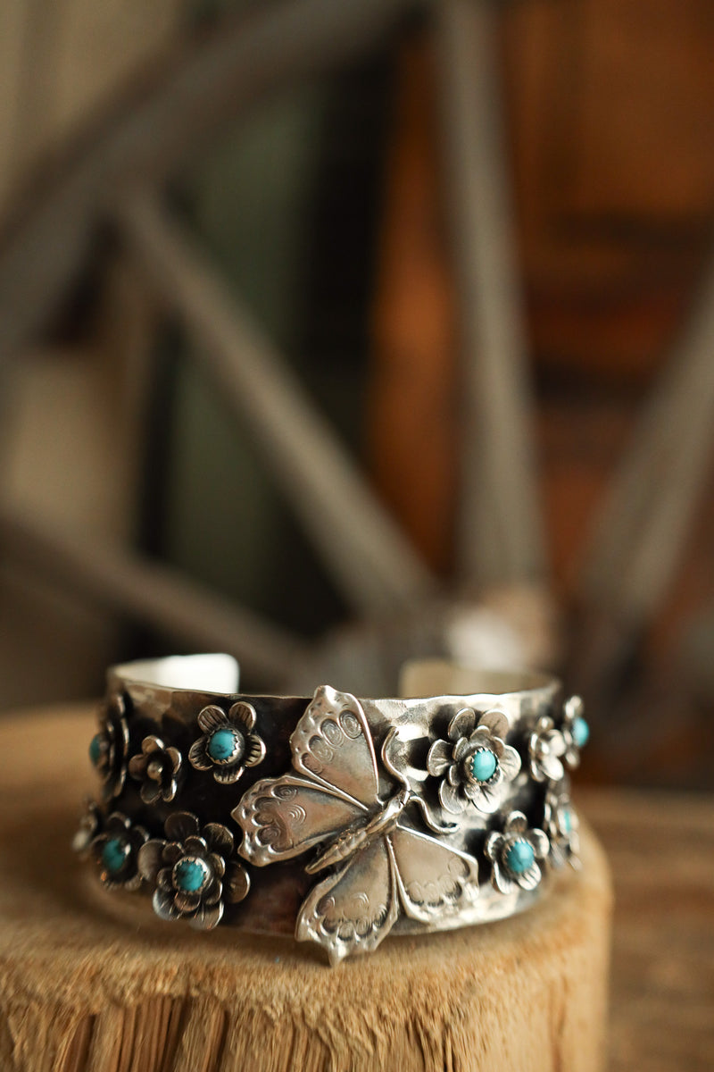 Silver bracelet with floral designs and turquoise stones on a wooden stand.