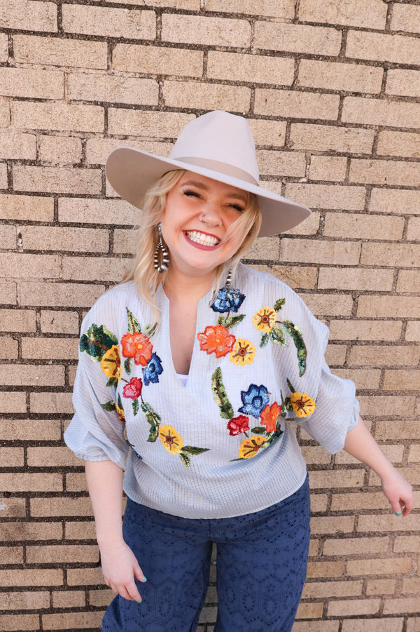 Woman wearing oversized blouse features a stunning blue checker print, a flirty v-neck, and chic quarter sleeve cinch detail. Adorned with multi-colored sequin flowers, this blouse will elevate any outfit with a touch of whimsy.