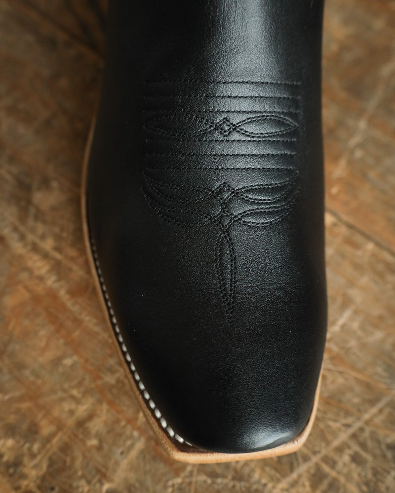 Close-up of a black leather boot with intricate stitching on a wooden surface