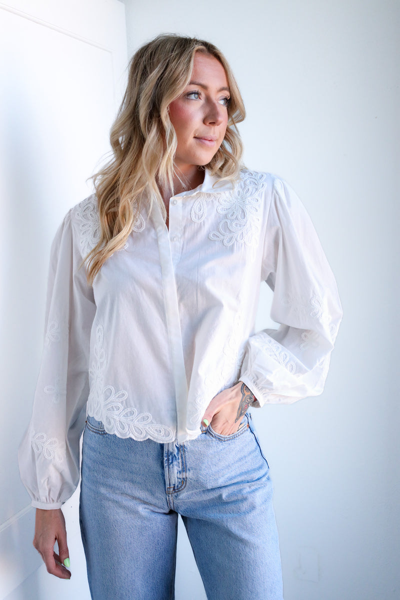 Woman wearing a white blouse with lace details and light blue jeans against a plain background