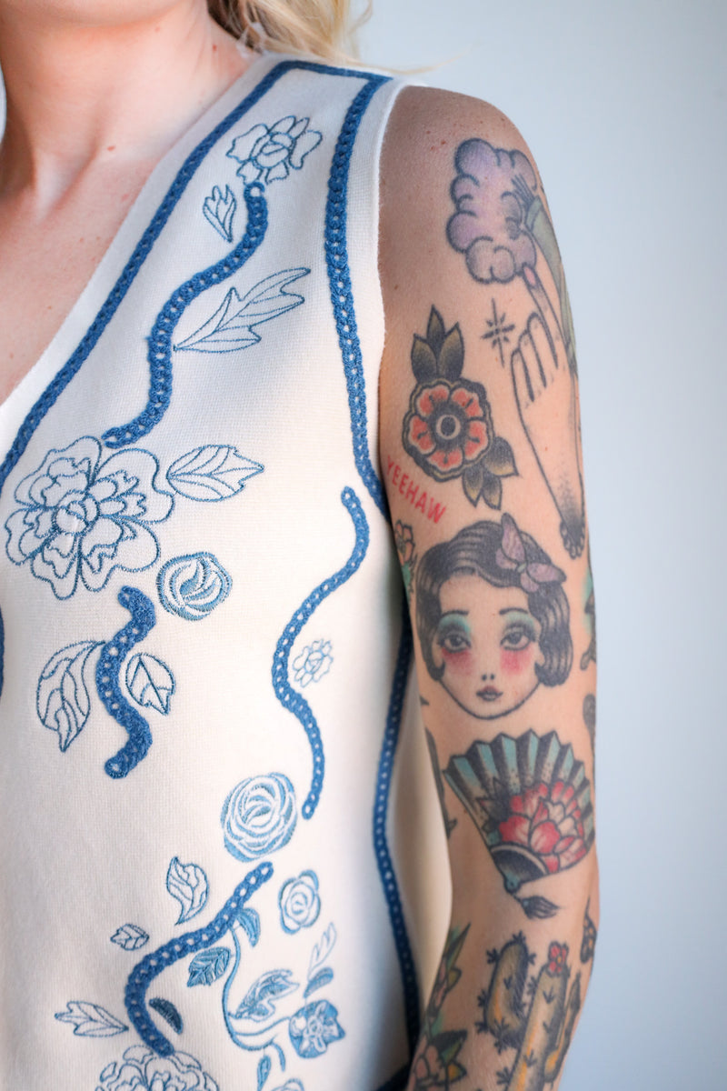 Person wearing a sleeveless top with blue floral embroidery and tattoos on the arm.