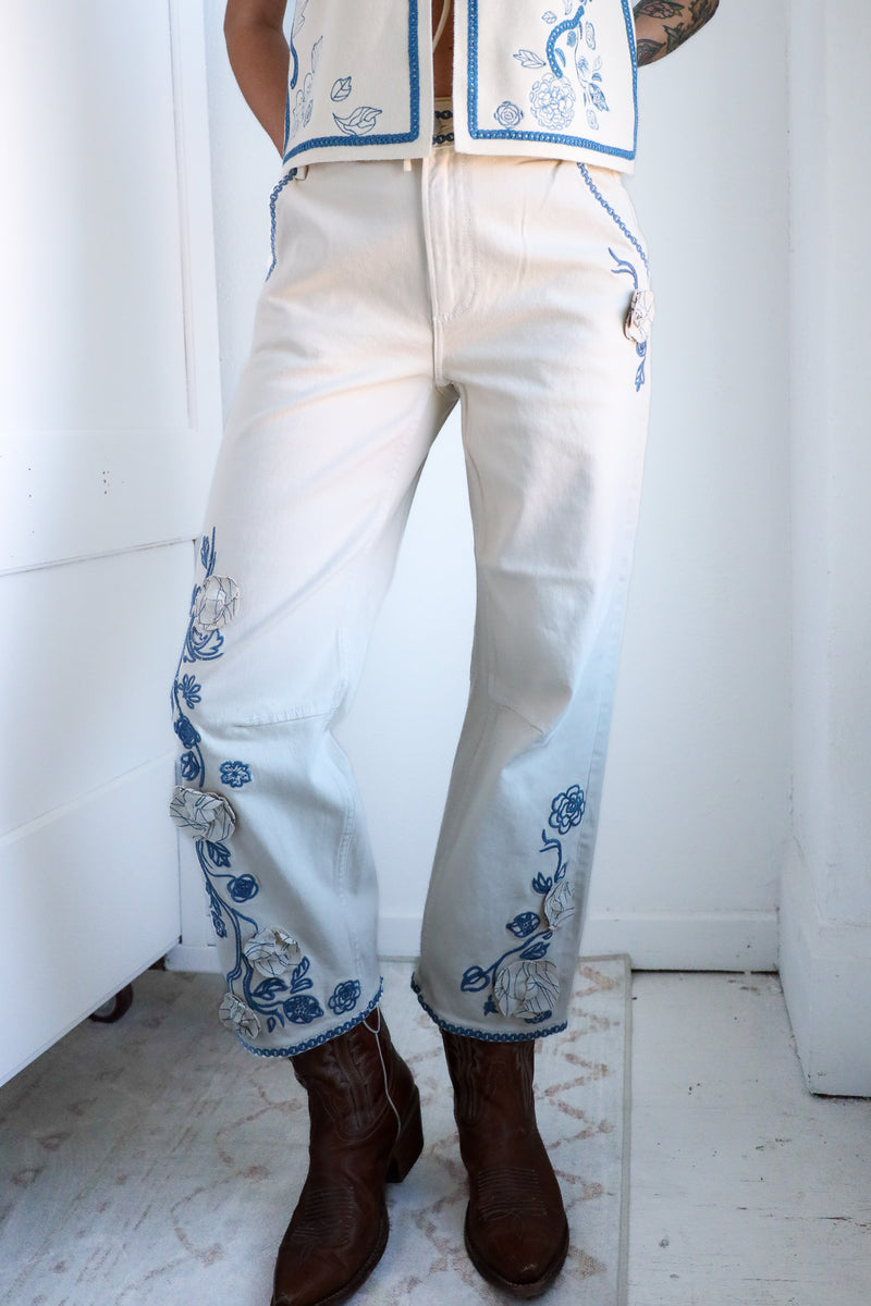 Person wearing white pants with blue floral embroidery and brown boots in a minimal indoor setting.
