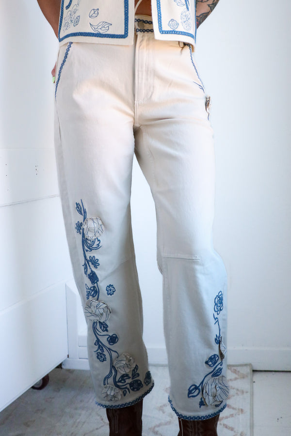 White pants with blue floral embroidery worn by a person against a white wall.