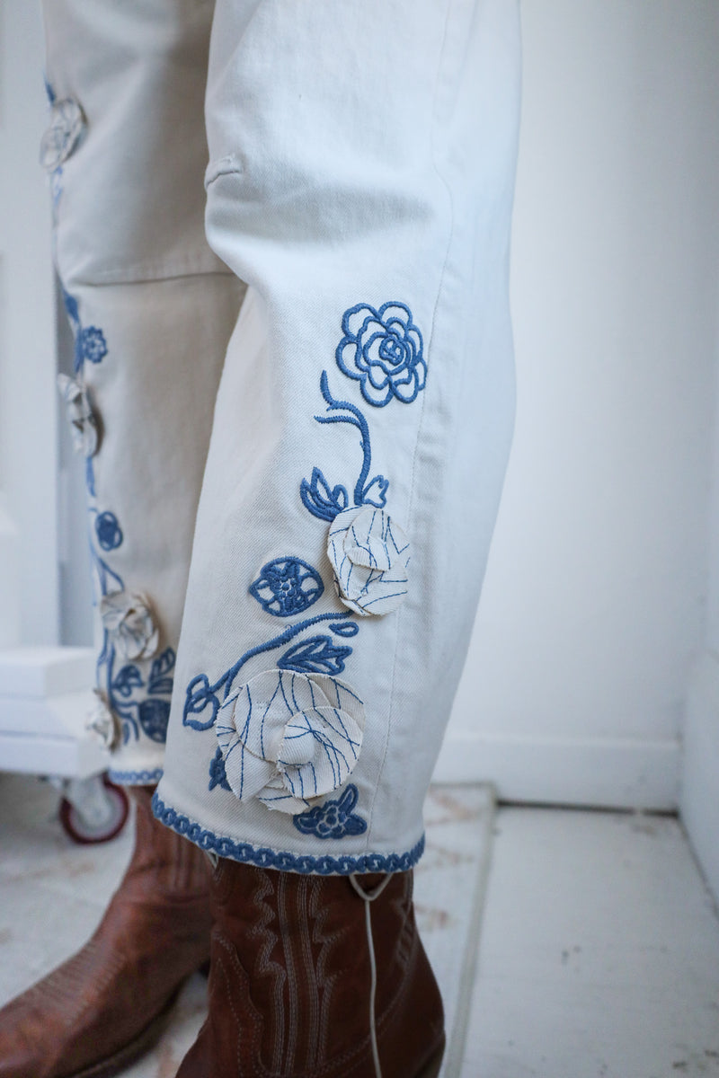 Close-up of white pants with blue floral embroidery and brown boots on a white background