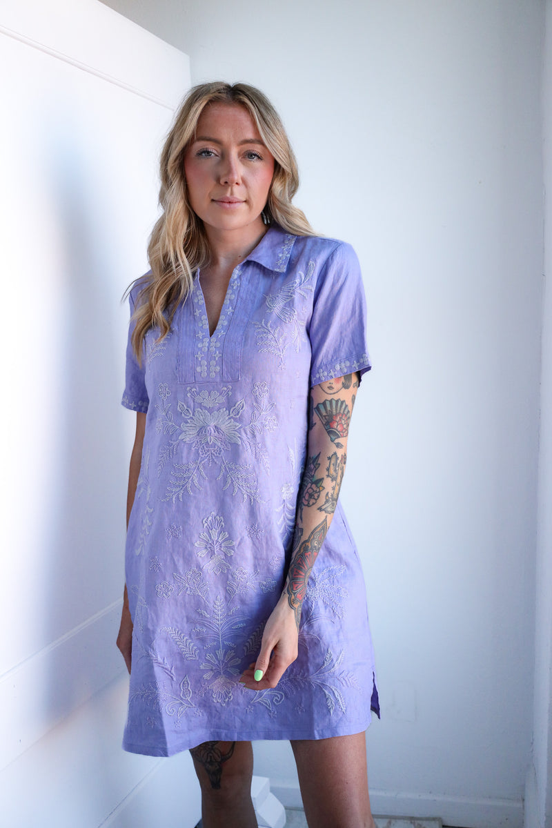 Woman wearing a light purple dress with lace details against a white wall.