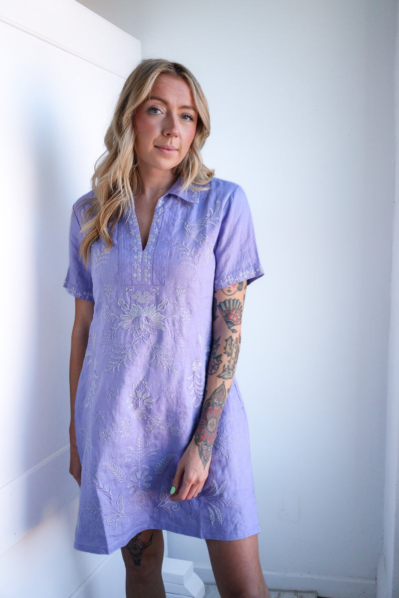 Woman wearing a purple dress with white patterns against a white wall.