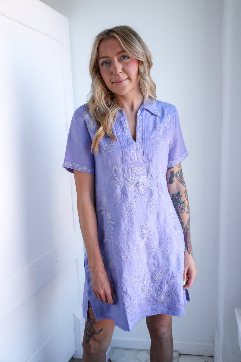 Woman wearing a light purple dress with lace details against a white wall.