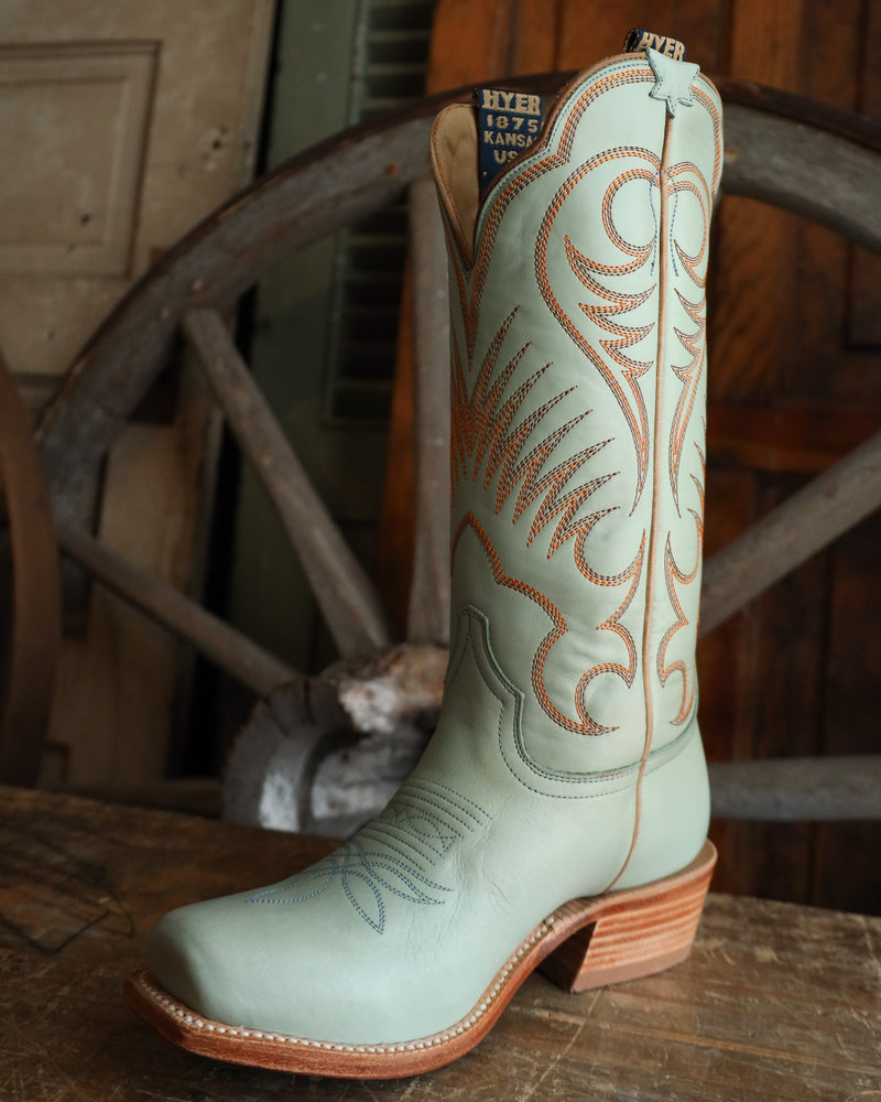 Light blue cowboy boot with brown stitching on a wooden surface with a wagon wheel in the background